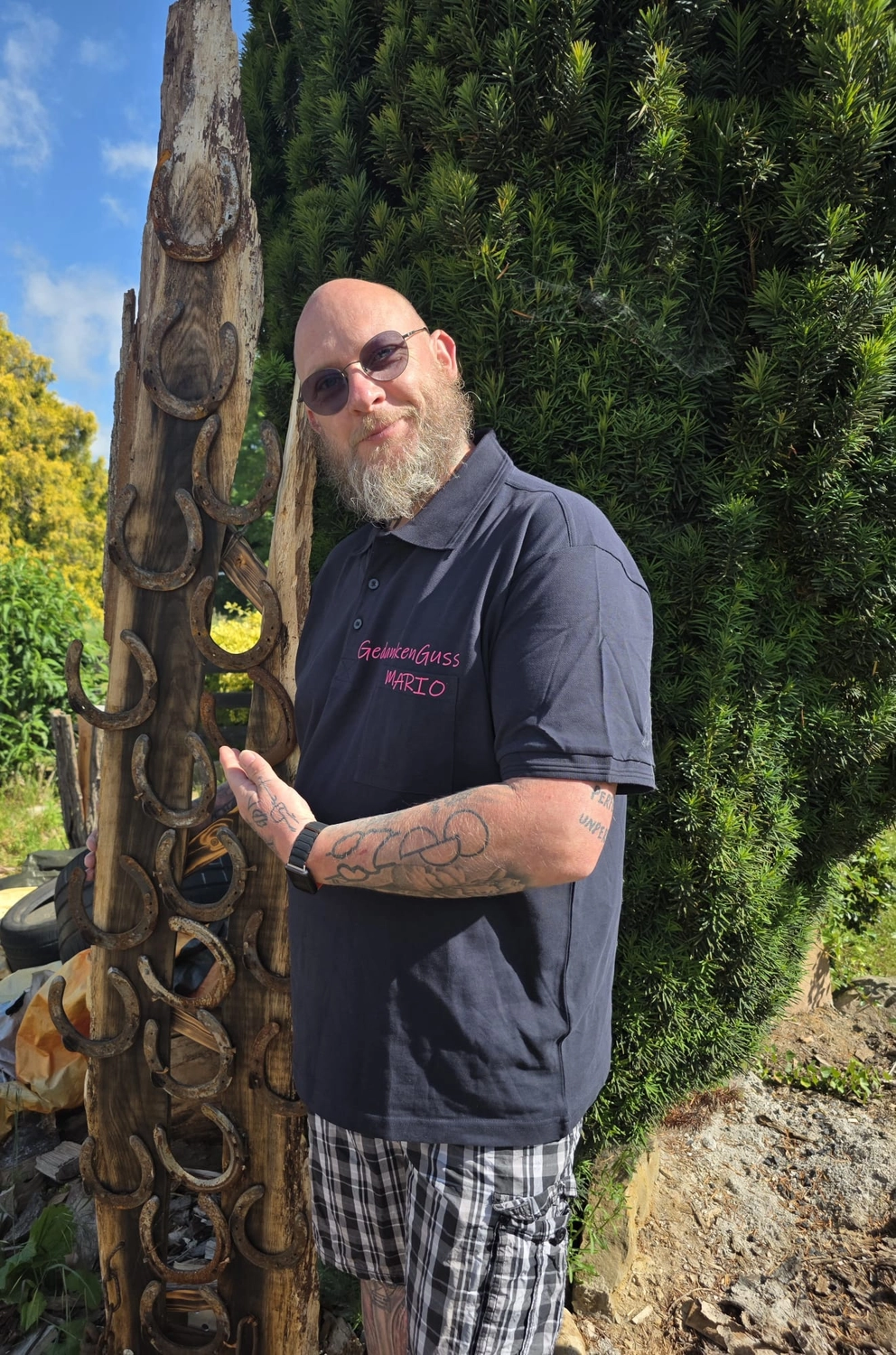 GedankenGuss - Holzskulpturen und abstrakten Kunstwerken - Mario Meinshausen mit Bart und Sonnenbrille steht neben einer mit Hufeisen verzierten Holzskulptur.